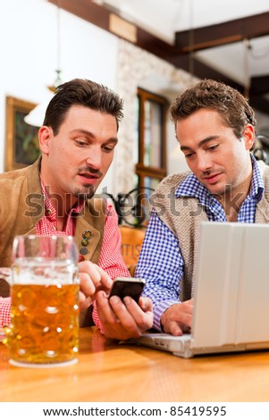 Two friends in Bavarian pub in traditional clothes sitting on a table in a pub with laptop