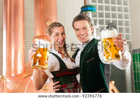 Man and woman with beer glasses in Bavarian tracht in brewery in front of a brew kettle