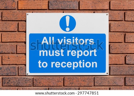 Mandatory all visitors must report to reception sign on a red brick wall outside a commercial building