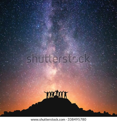 Together we stand. A group of people are standing on the top of the hill next to the Milky Way galaxy holding hands.