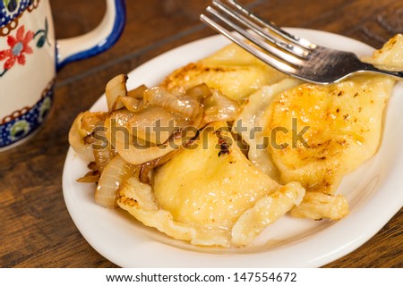 A plate of handmade pierogi with a side of carmelized onions.  There is a silver fork on the plate, as well as a colorful mug in the background.  Everything is shot on a wood table.
