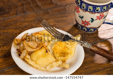 A plate of handmade pierogi with a side of caramelized onions. There's a decorative silver and wood fork on the plate, as well as a colorful mug in the background.  Everything is shot on a wood table.