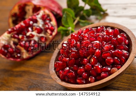 Loose pomegranate (Punica granatum) seeds in a wood bowl shot on a wood table. There are pieces of ripe pomegranate fruit & green mint leaves in the background. Pomegranates are a super food.