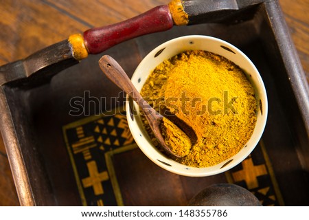 Ceramic bowl filled with yellow curry powder with a wood spoon on a decorative wooden tray. The tray is shot on a wood surface.