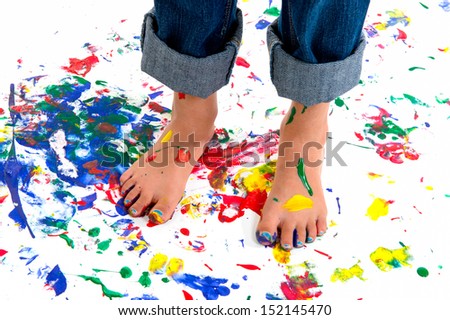 A young girl\'s feet covered in colorful paint on a paint splashed background.  She has her jeans rolled up to avoid the mess.