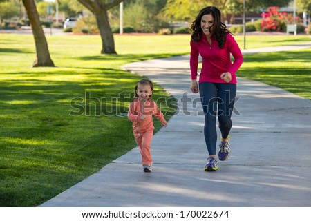 Beautiful young mother and her daughter running in the neighborhood.  They are on a sidewalk in a grassy greenbelt.  The mother is playfully chasing her daughter.