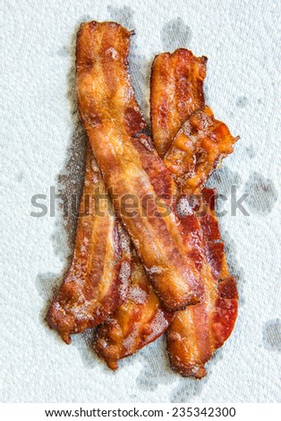 Strips of cooked bacon piled up on a white paper towel.  You can see all of the grease being absorbed by the paper towel.