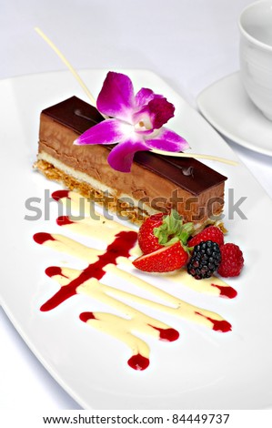 Beautifully plated chocolate mousse cake garnished with a purple orchid, strawberry, blackberry and raspberry.  A zigzag of white chocolate and strawberry sauce completes the plate.