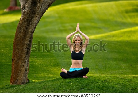 Beautiful blond young woman practicing yoga in the park on a green lawn. Her arms are together over her head and she is sitting on the group with her legs crossed.
