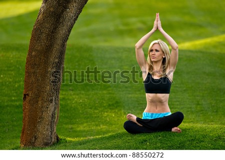 Beautiful blond young woman practicing yoga in the park on a green lawn. Her arms are together over her head.