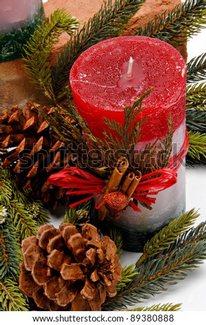 Red, white and green festive candle with green cedar leaves and cinnamon sticks tied on with twine for Christmas. The candle is placed among pine branches and pine cones.