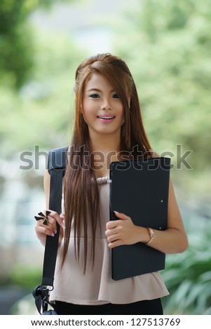 Young woman in business attire, carrying briefcase and holding folder standing outside.