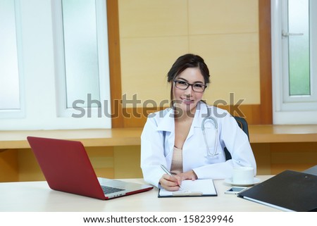 Attractive young female doctor sitting at desk in office doing paperwork, looking up and smiling at camera.