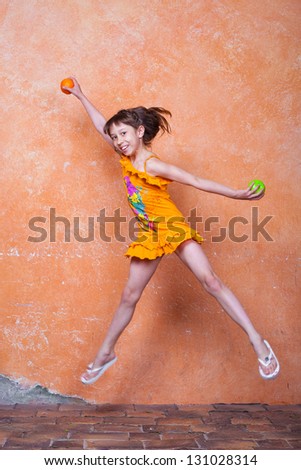 little girl in orange dress jumping beside the orange wall with orange and apple in her hands