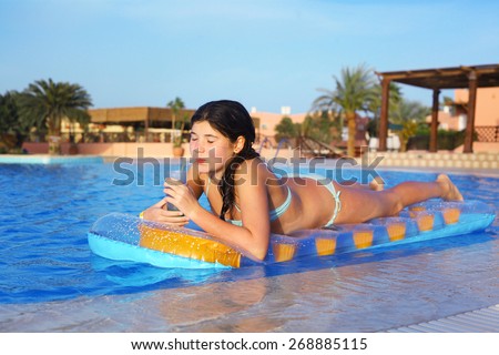 preteen girl in swimming suit with inflatable matrass drink cocktail on the blue pool water and palms background