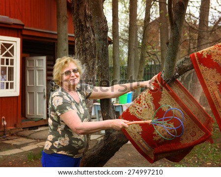 happy pensioner woman grandma beat red rugs in the garden on the mansion house background