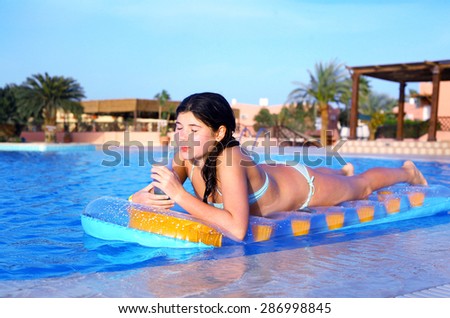 preteen girl in swimming suit with inflatable matrass drink cocktail on the blue pool water and palms background