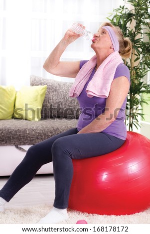 senior woman sitting on a gym ball at home after training and drink water