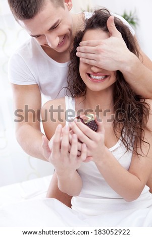 Handsome young man sitting on the bed and holding a gift box  covering his girlfriend  eyes with hands