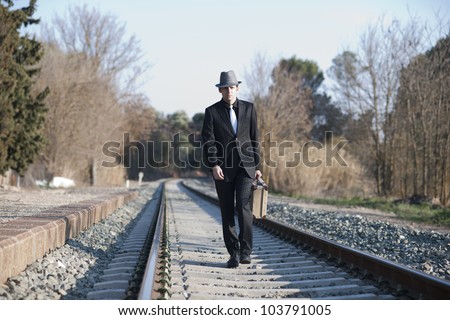 Man with suit , hat and suitcase walking across the railway