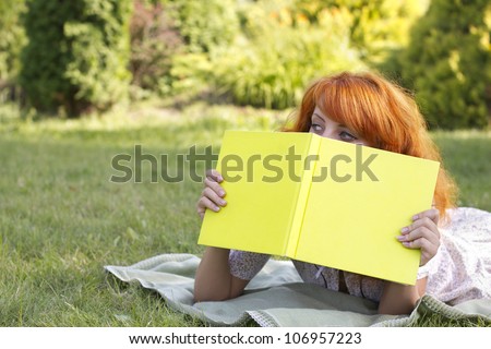 Redhead woman lies on green grass and reads green  book, covers the face during spring / summer time . Happy smiling beautiful young university student studying lying down in grass.