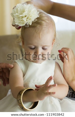 Little, blond baby girl getting ready for the celebration and dressing up in jewelry. Baby dressed in a white fluffy dress, bracelet, bow on her head. Dress up