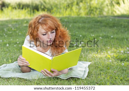 Redhead woman lies on green grass and reads green book, covers the face during spring / summer time . Happy smiling beautiful young university student studying lying down in grass.