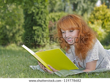 Redhead woman lies on green grass and reads green book, covers the face during spring / summer time . Happy smiling beautiful young university student studying lying down in grass.
