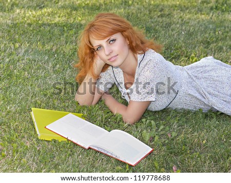 Redhead woman lies on green grass and reads green book, covers the face during spring / summer time . Happy smiling beautiful young university student studying lying down in grass with.