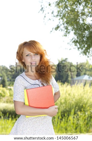 Redhead woman standing in park near university, college with books on nature in summer or spring. Happy smiling beautiful young girl university student