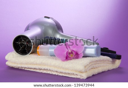 The hair dryer, hairbrush, shampoo on a towel, decorated with a flower, on the violet