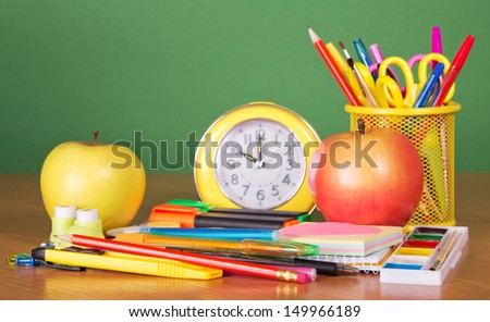 Pencils, handles, scissors in a support, an alarm clock and two apples, on a green background