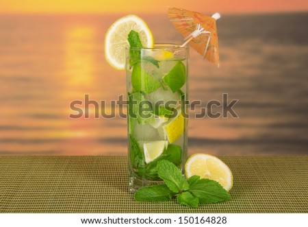 Cold drink with a lemon slice, spearmint, on a green bamboo cloth against the sunset