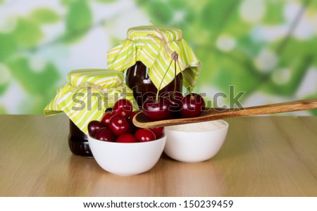 Two jars of jam, cup of sugar, wooden spoon with cherries, berries in a cup on a table