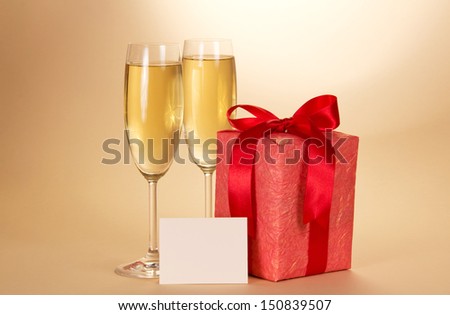 Two wine glasses with champagne, a gift box with a bow, an empty card on a beige background
