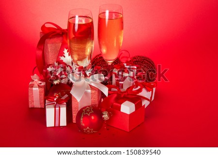 Two wine glasses with champagne, gift boxes with bows and tapes, Christmas spheres and serpentine on a red background