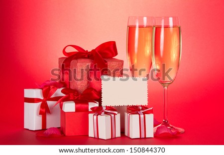 Two glasses of champagne, gift boxes with tapes and bows, empty card and petals of flowers on a red background