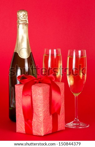 Gift box with a tape and bow, bottle of champagne and the wine glasses on a red background