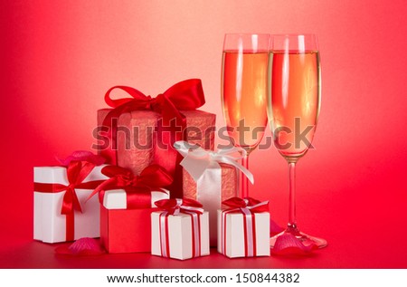 Two glasses of champagne, gift boxes with tapes and bows and petals of flowers on a red background