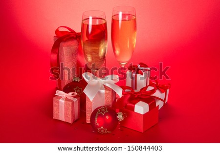 Two wine glasses with champagne, gift boxes with bows and tapes and Christmas sphere on a red background