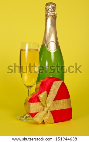 Bottle and wine glass with champagne, a gift box heart on a yellow background