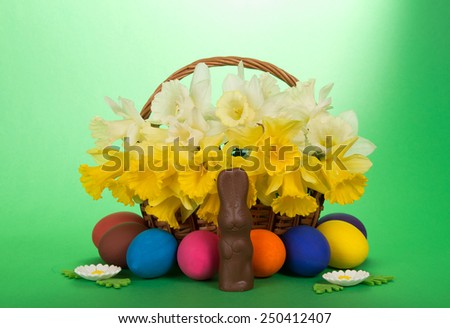 White and yellow narcissuses in a wattled basket, eggs, flowers and a chocolate rabbit, on a green background