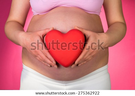 Pregnant woman holding red heart near her belly, close up on pink background