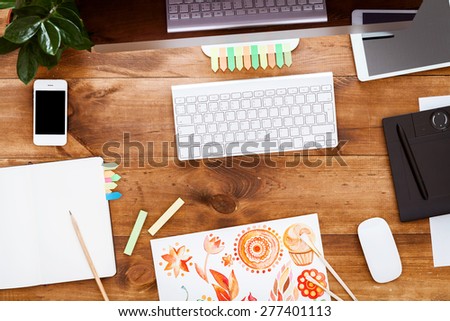 Brown wooden table, top view. On the table are, stationery items, notebook, gadgets, green flower, stickers, keyboard, mouse, a sheet of paper with a picture, crayons.