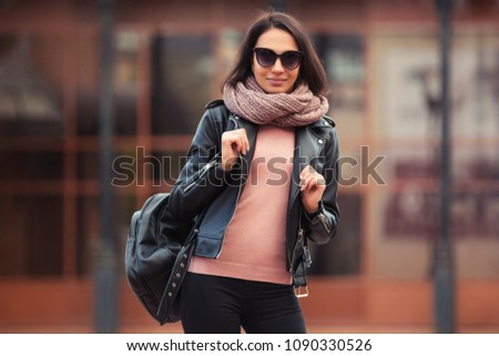 Young fashion woman with backpack walking on city street Stylish female model in black leather jacket and snood scarf