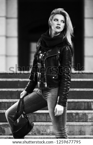 Young fashion blonde woman with handbag walking on city street  Stylish female model wearing black leather jacket and scarf