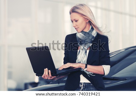 Young fashion business woman with laptop next to her car on parking. Stylish female model in black jacket and blue scarf outdoor