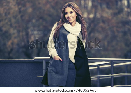 Happy young woman walking on city street Stylish fashion model in long sleeveless coat and white sweater