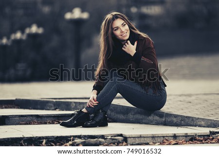 Happy young woman sitting on the sidewalk on city street Stylish fashion model in a fringe leather suede jacket and dark blue jeans