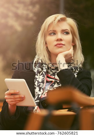 Young blond business woman using tablet computer sitting on the bench. Stylish fashion female model in black coat in a city street